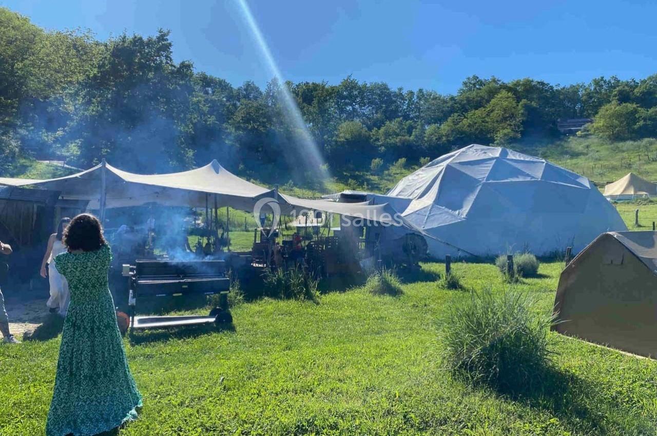 Femme en robe verte près d'un barbecue fumant, avec des tentes et une structure géodésique dans un paysage verdoyant.