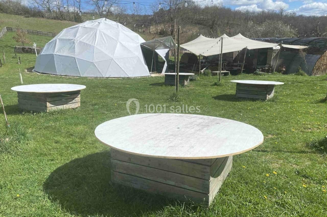 Tables rondes en bois disposées sur une pelouse, avec une structure géodésique blanche et des tentes en arrière-plan.