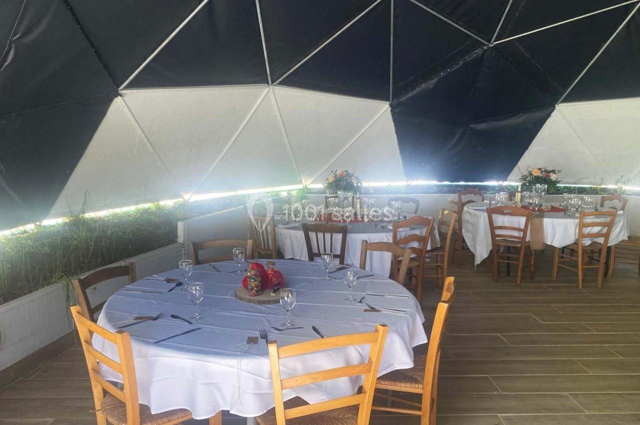 Salle sous dôme géodésique avec tables rondes dressées, nappes blanches, chaises en bois et décorations florales.