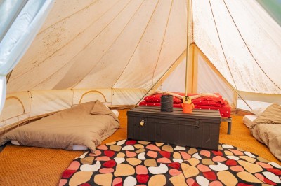 Intérieur d'une tente avec matelas, coffre noir, tapis coloré et vue partielle sur un paysage extérieur.