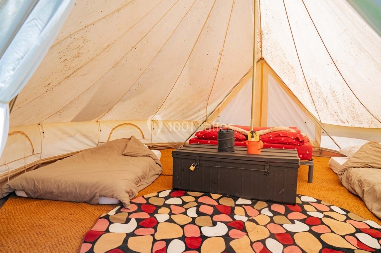 Intérieur d'une tente avec lits simples, coffre en bois, tapis coloré et lumière naturelle filtrant à travers la toile.