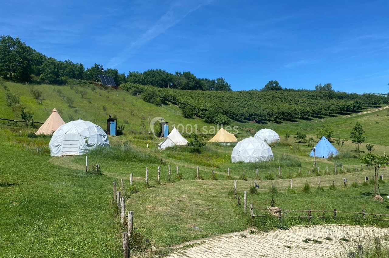 Plusieurs tentes et dômes géodésiques installés dans un paysage vallonné avec des arbres et un ciel dégagé.