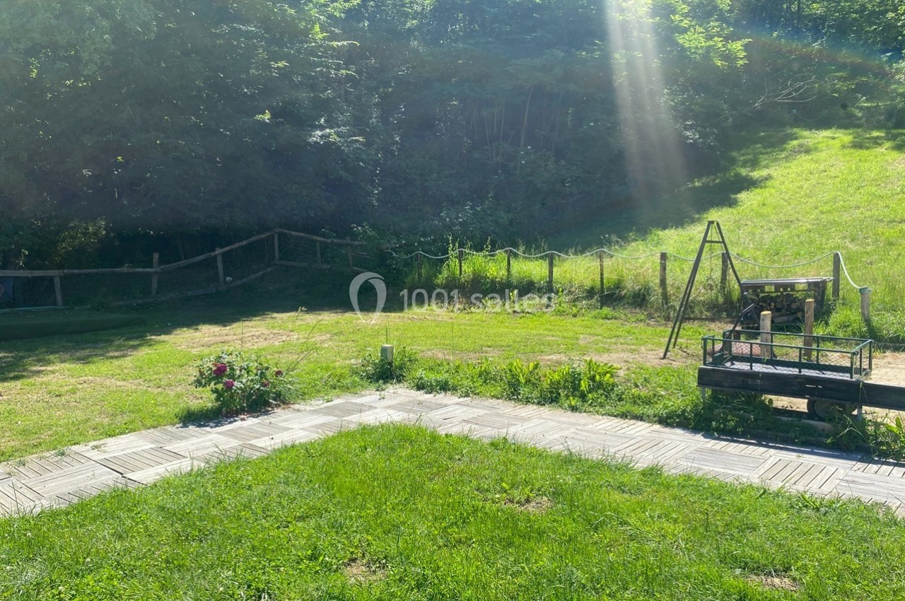 Jardin ensoleillé avec pelouse, allée en dalles, clôture en bois, balançoire et arbres en arrière-plan.