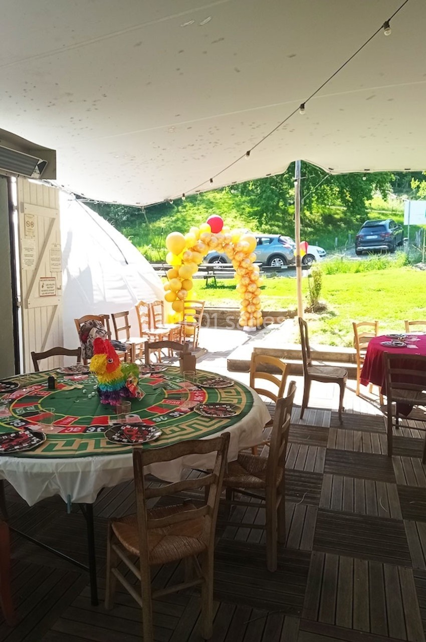 Salle extérieure décorée avec des tables rondes, nappes colorées et arche de ballons jaunes sous un auvent.