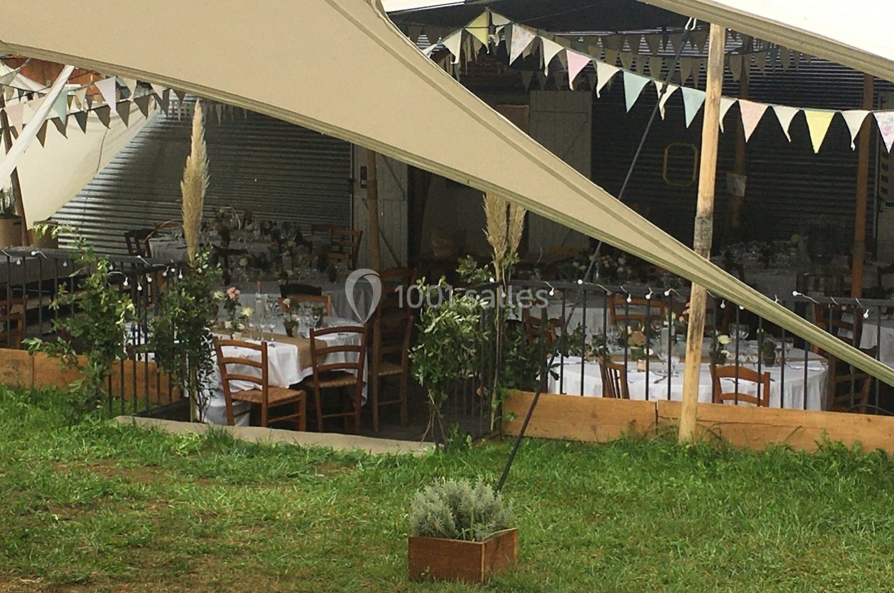 Tables dressées avec nappes blanches sous une tente, décorées de guirlandes et de végétation, dans un cadre extérieur.