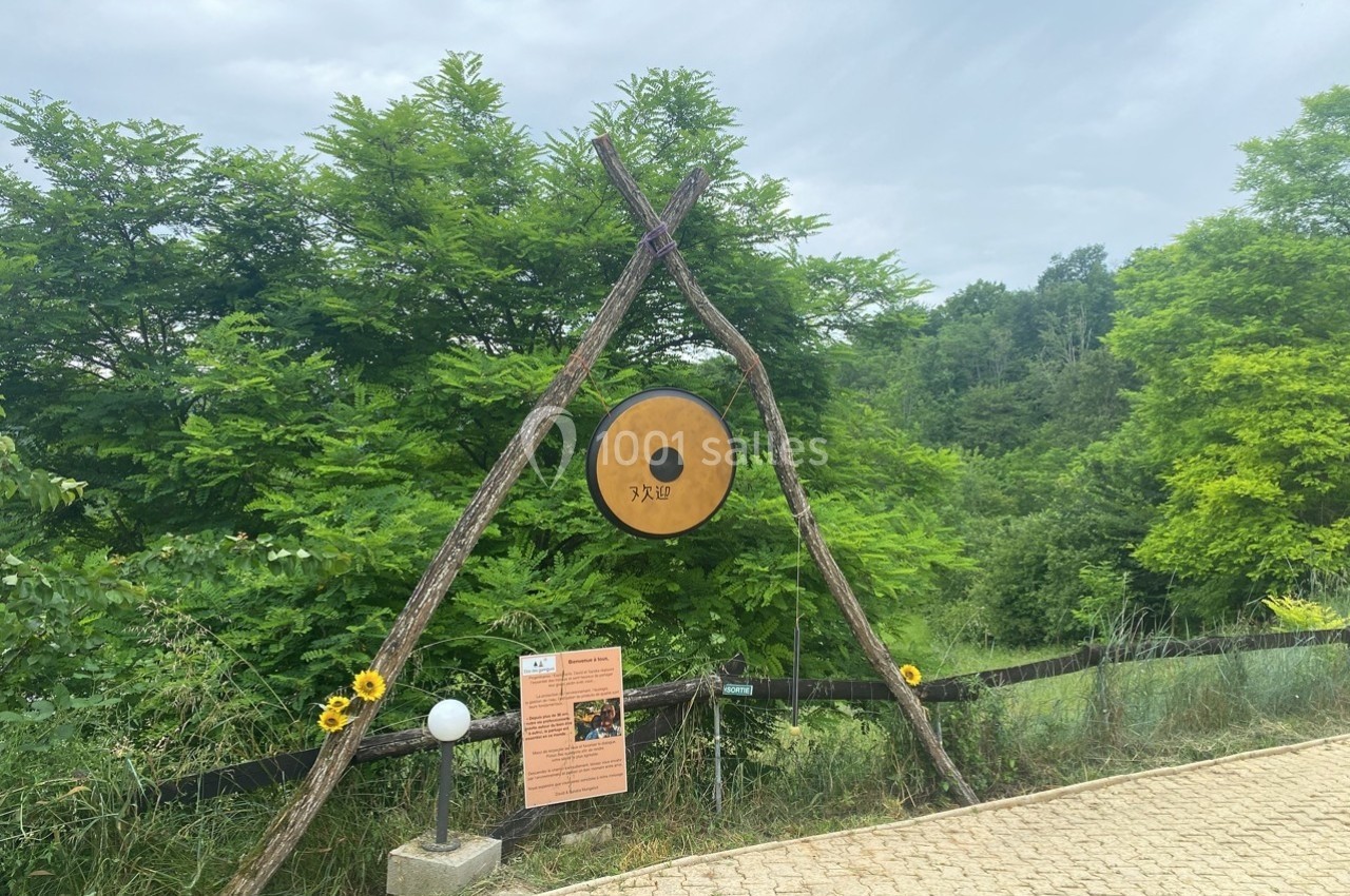Un grand gong suspendu entre deux branches en triangle, entouré de verdure, près d'un chemin pavé.