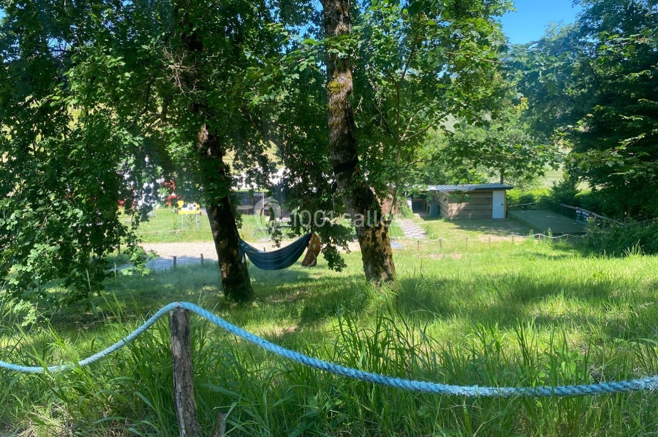 Hamac suspendu entre deux arbres dans un espace verdoyant, avec des bâtiments en arrière-plan.