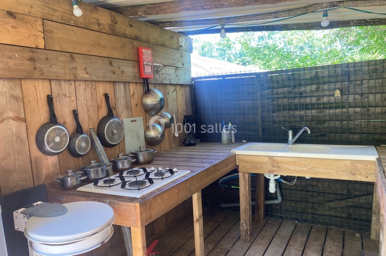Cuisine extérieure en bois avec évier, plaques de cuisson, casseroles suspendues et extincteur fixé au mur.
