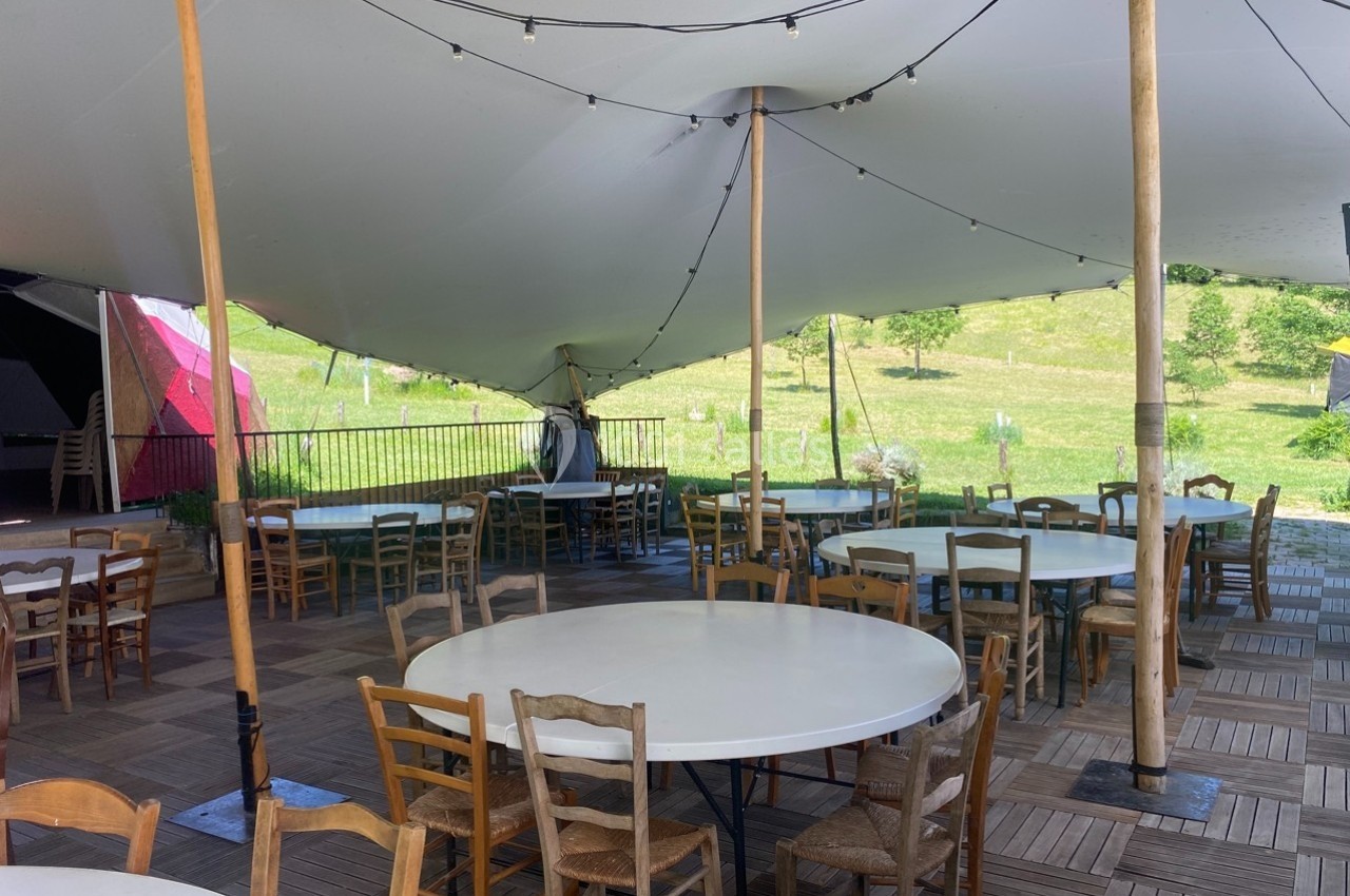 Tables rondes et chaises en bois disposées sous une grande tente blanche dans un espace extérieur avec vue sur la campagne.