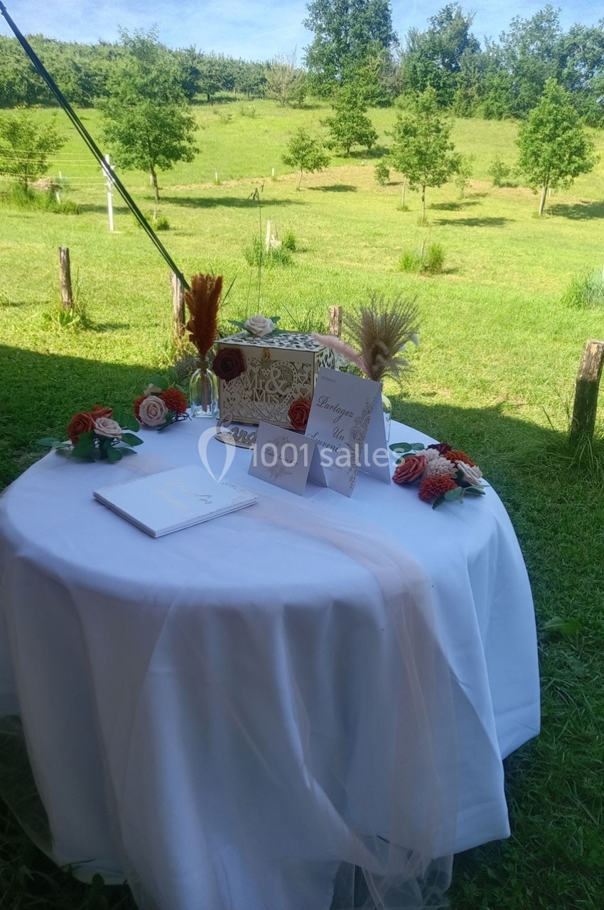 Table ronde décorée pour un mariage, avec urne, fleurs, cartes et vue sur un jardin verdoyant en arrière-plan.