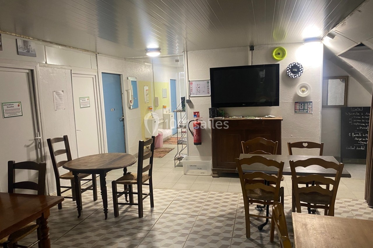 Salle commune avec tables, chaises, télévision murale, extincteur et décor sobre dans un espace lumineux.