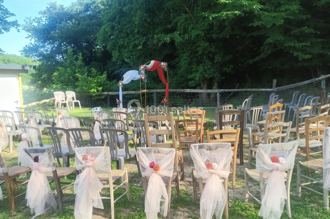 Chaises dépareillées décorées de tulle et de fleurs, disposées en extérieur devant une arche ornée de tissu rouge et blanc.