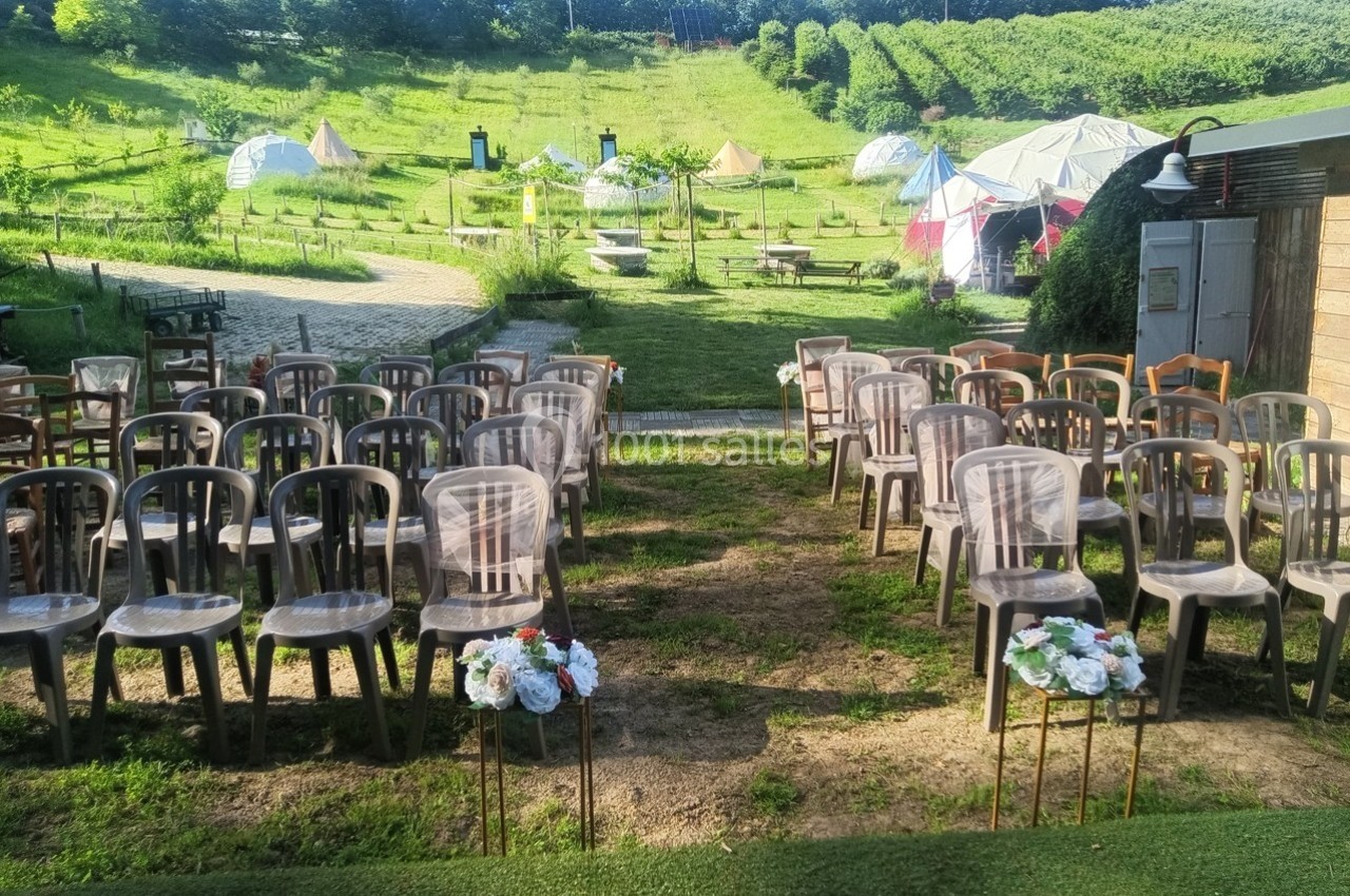 Chaises disposées en rangées en extérieur, avec des décorations florales, devant un paysage verdoyant et des tentes.