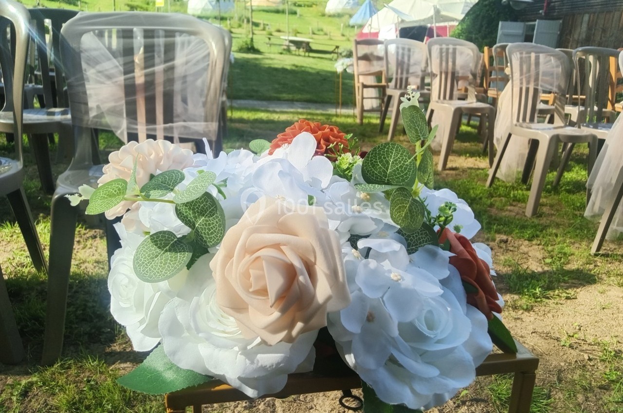 Arrangement de fleurs blanches, beiges et orange sur une table, avec des chaises disposées en extérieur en arrière-plan.