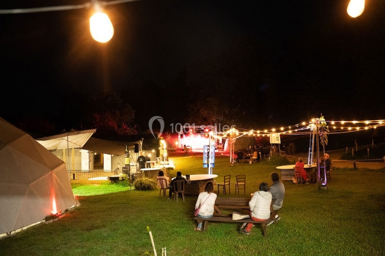 Espace extérieur éclairé de nuit avec des guirlandes lumineuses, des structures et des personnes assises sur des bancs.