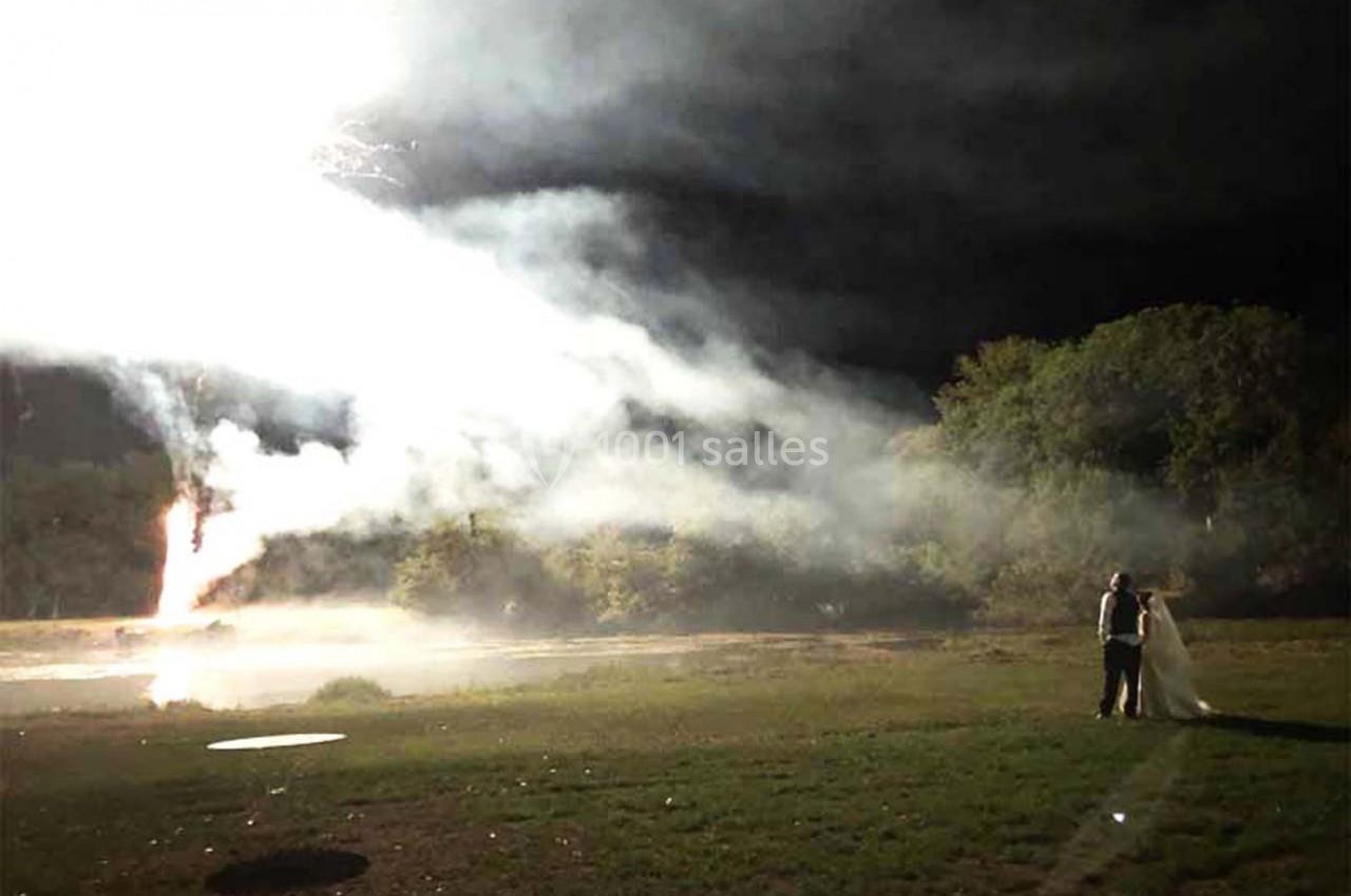 Un couple debout sur une pelouse regarde un feu d'artifice illuminant un ciel nocturne avec de la fumée.