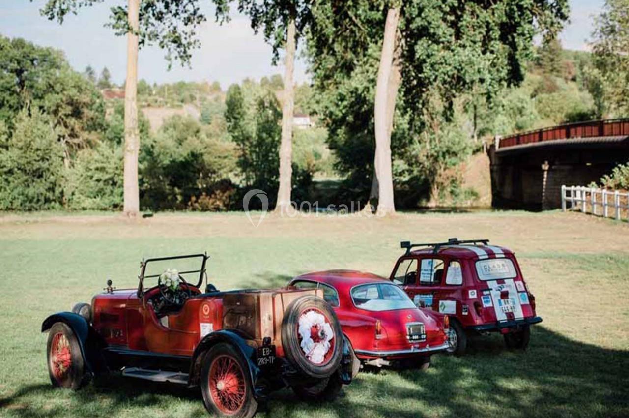 Trois voitures anciennes rouges stationnées sur une pelouse, entourées d'arbres et d'un pont en arrière-plan.