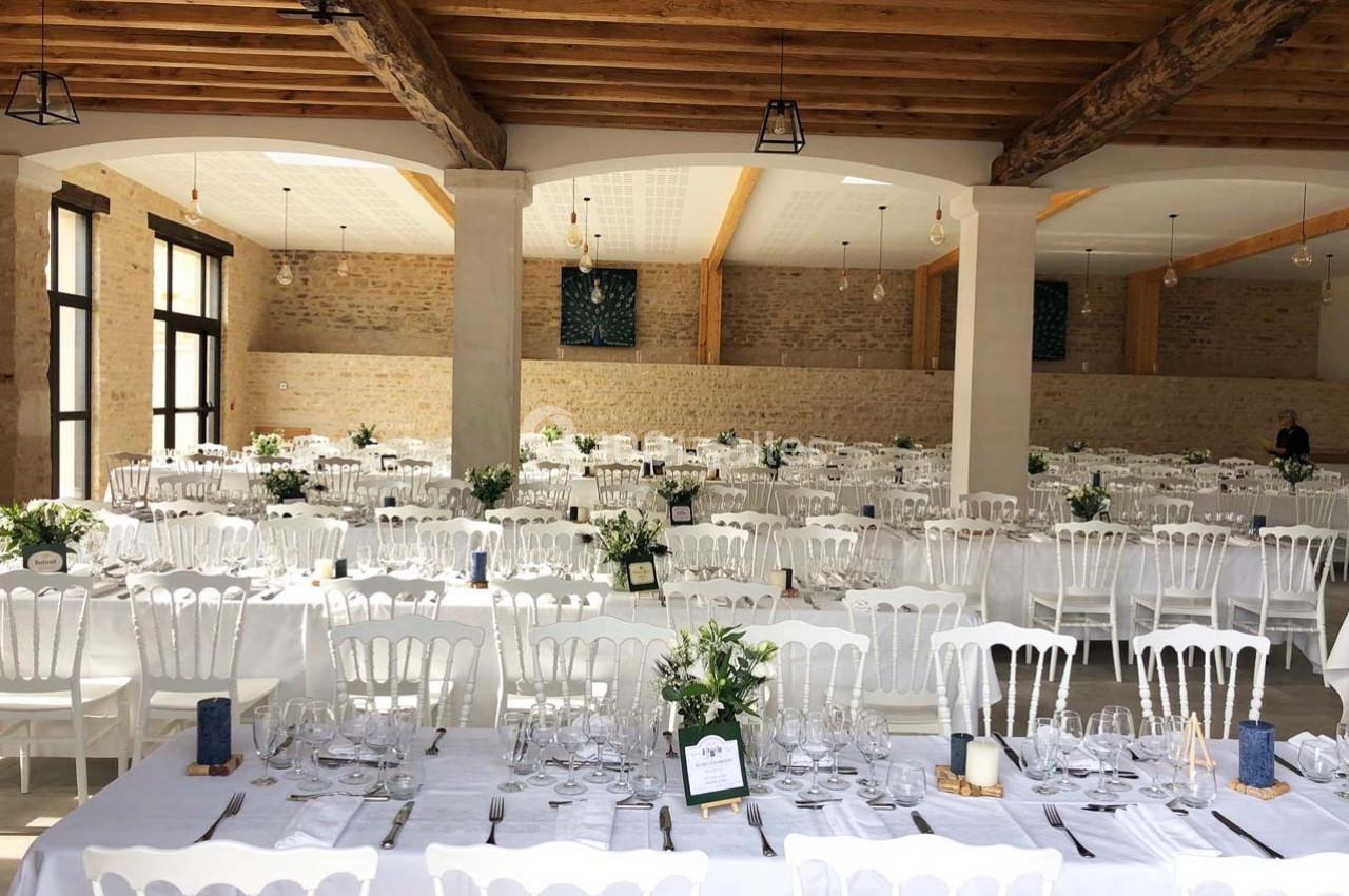 Salle de réception lumineuse avec de longues tables dressées, chaises blanches et décorations florales.