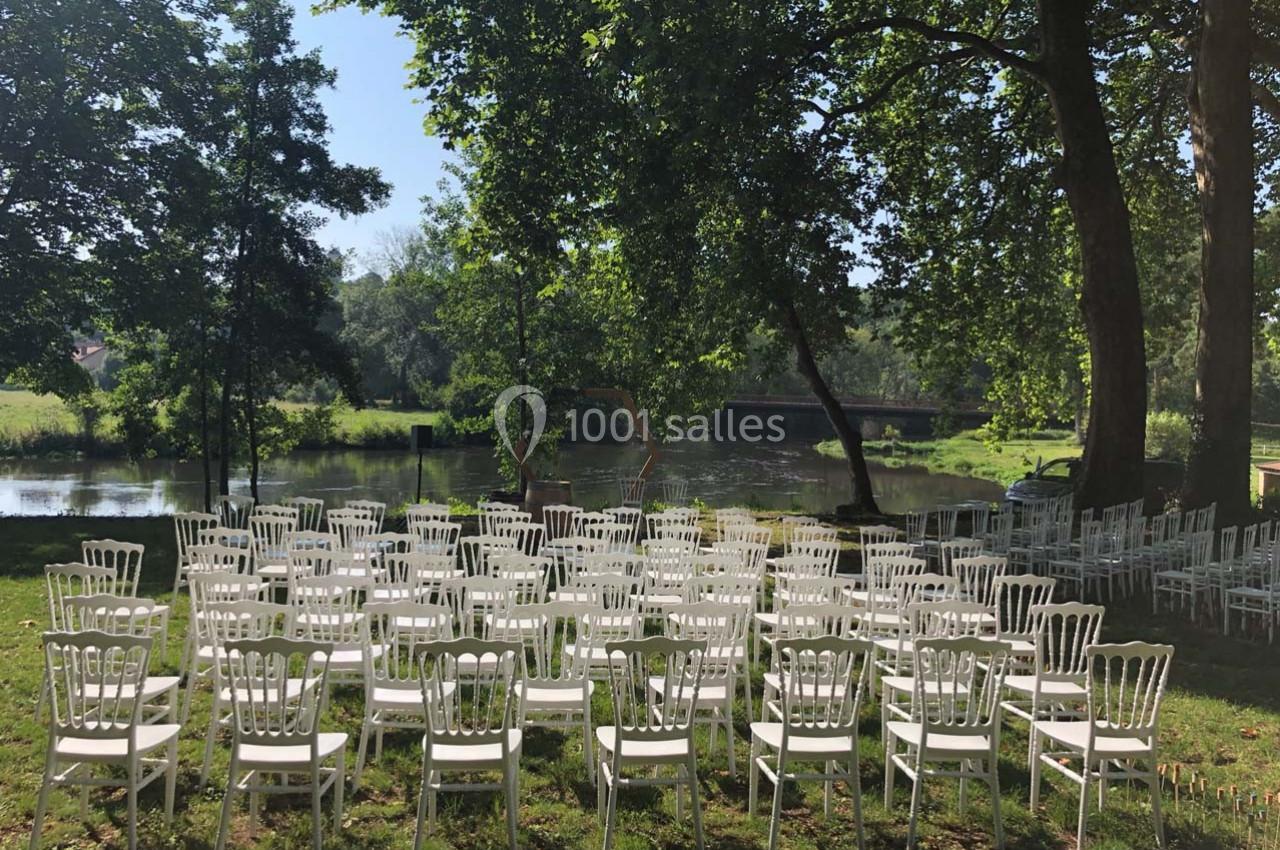 Chaises blanches disposées en rangées sur une pelouse, face à une rivière bordée d'arbres par une journée ensoleillée.