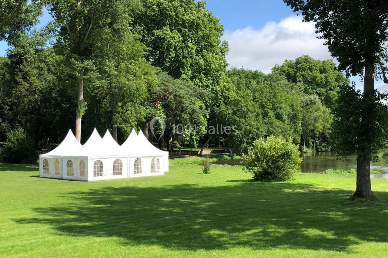 Tente blanche avec des fenêtres, installée sur une pelouse verdoyante près d'un plan d'eau entouré d'arbres.