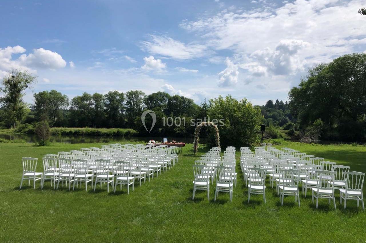 Chaises blanches disposées en rangées sur une pelouse, formant une allée centrale, avec une arche décorée en arrière-plan.