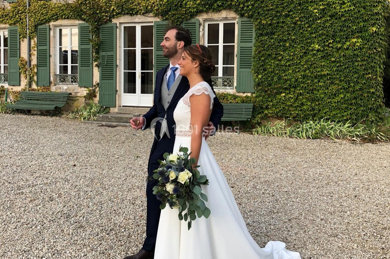 Un couple en tenue de mariage pose devant une maison en pierre couverte de lierre, sur une allée de gravier.