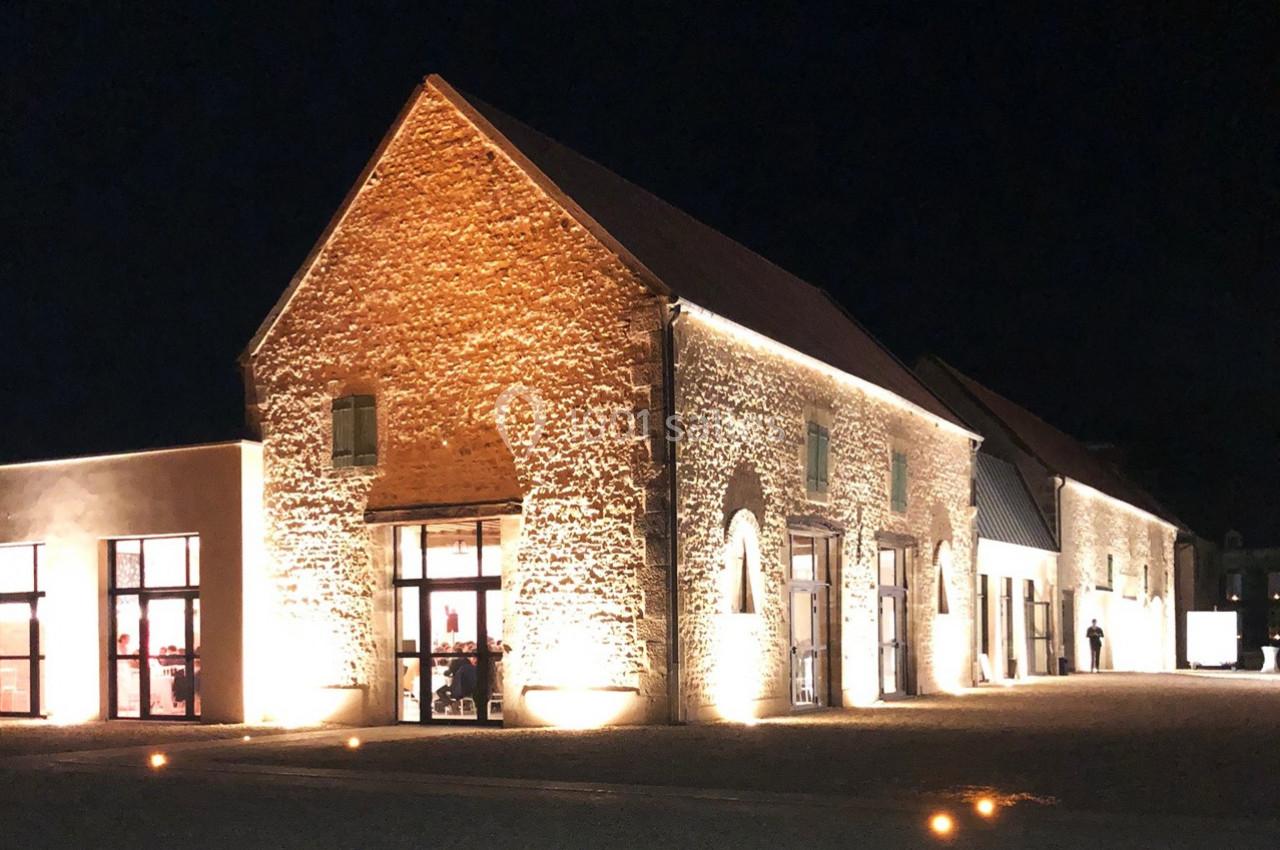 Bâtiment en pierre éclairé la nuit, avec grandes fenêtres illuminées et une cour devant.