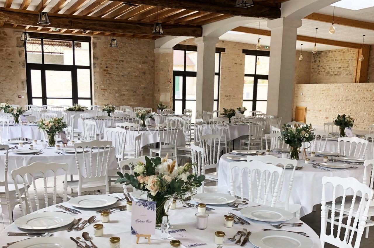 Salle de réception décorée avec des tables rondes, nappes blanches, chaises en bois blanc et bouquets floraux.