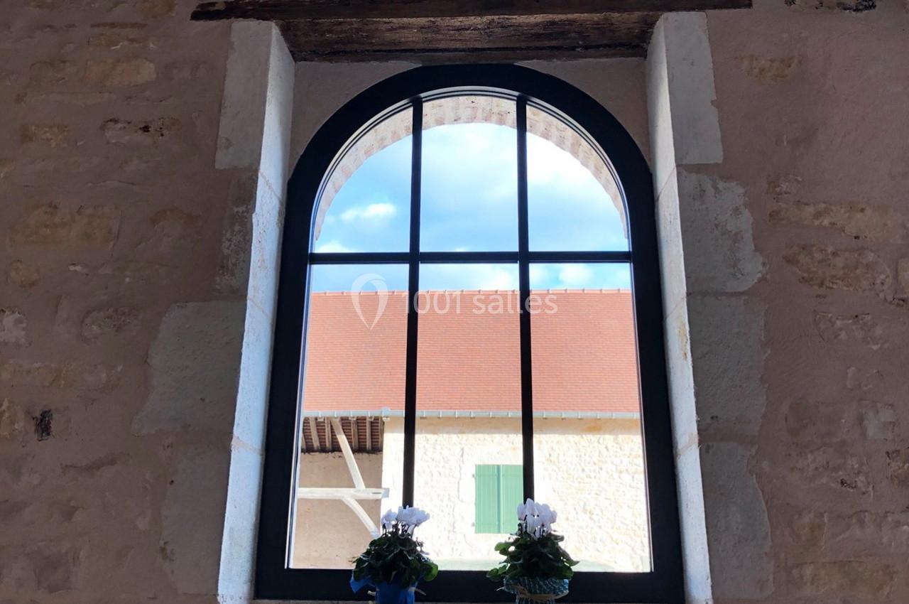 Fenêtre cintrée avec cadre noir, vue sur un toit en tuiles rouges et deux pots de fleurs blanches sur le rebord.