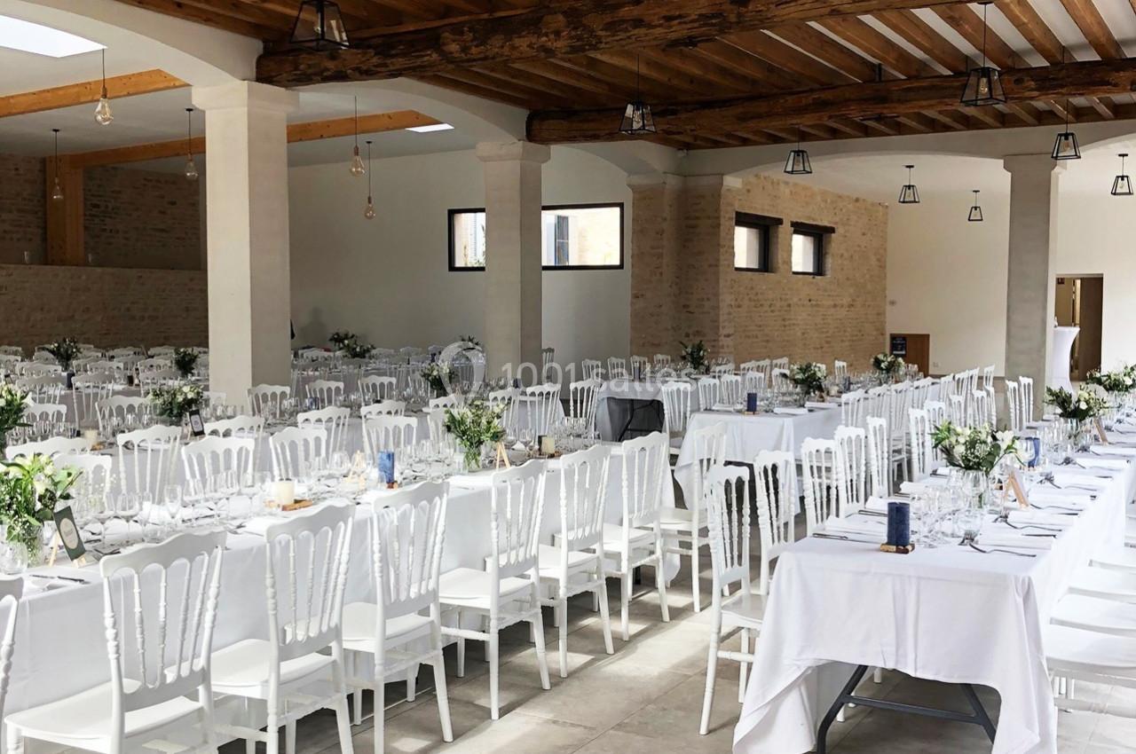 Salle de réception lumineuse avec de longues tables décorées de nappes blanches, chaises blanches et arrangements floraux.