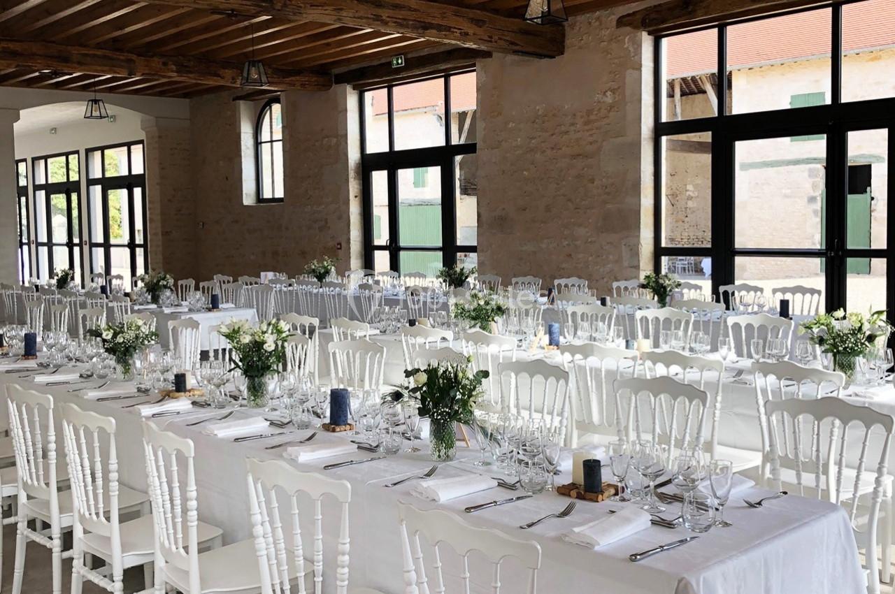 Salle de réception lumineuse avec de longues tables décorées de nappes blanches, fleurs et vaisselle élégante.