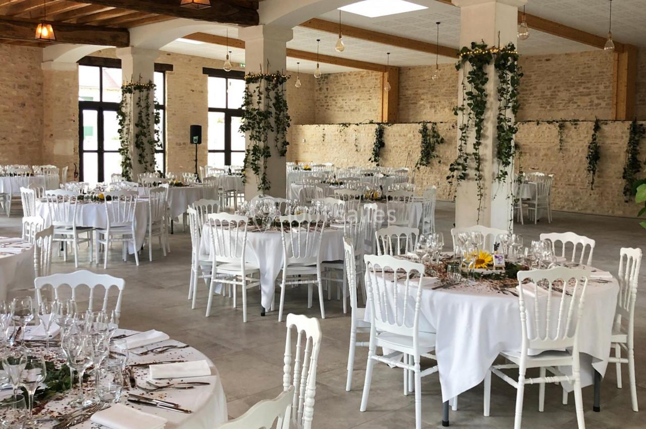 Salle de réception lumineuse avec tables rondes décorées, nappes blanches et chaises en bois, ornée de végétation suspendue.