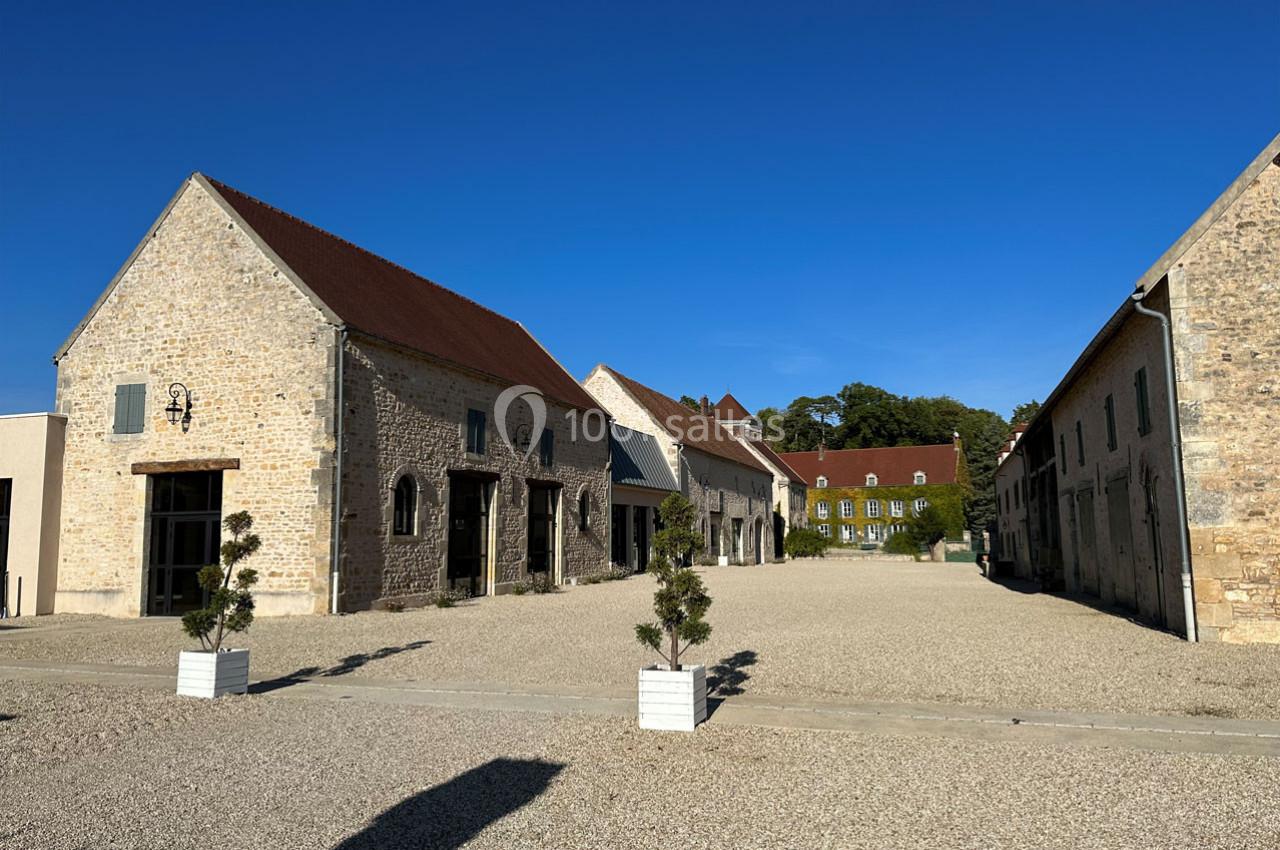 Bâtiments en pierre avec toits en tuiles rouges entourant une cour gravillonnée sous un ciel bleu clair.