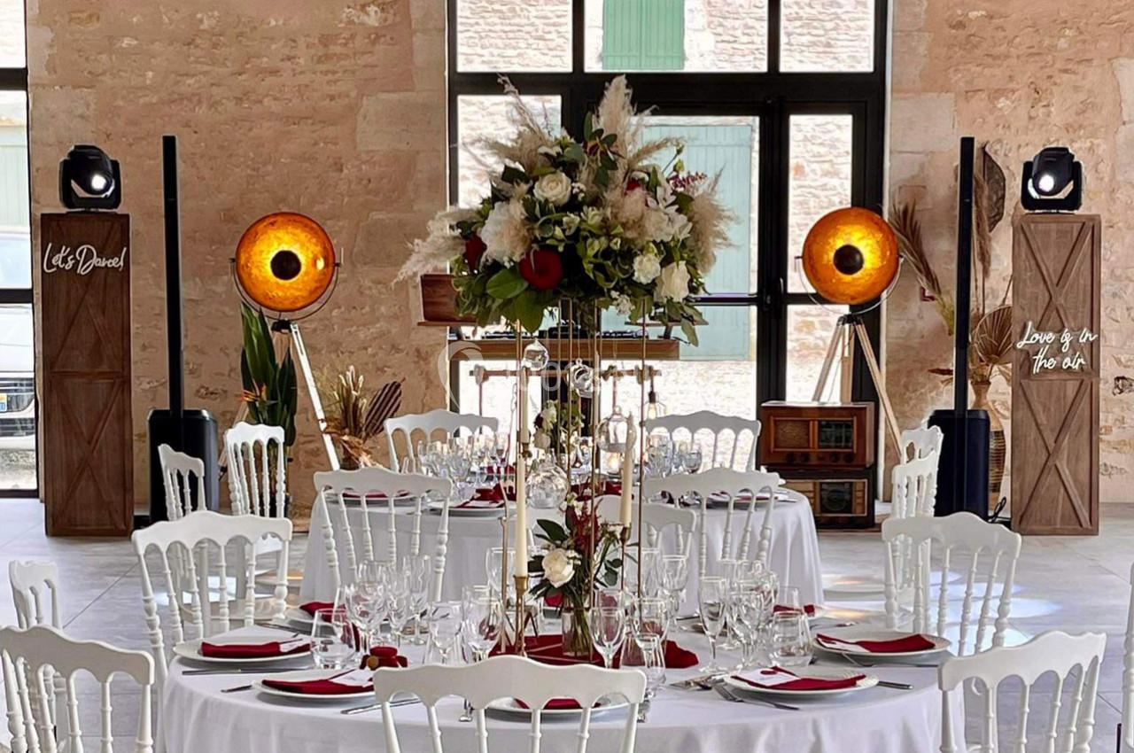 Salle de réception décorée avec des tables rondes, des chaises blanches, un centre de table floral et des éclairages…