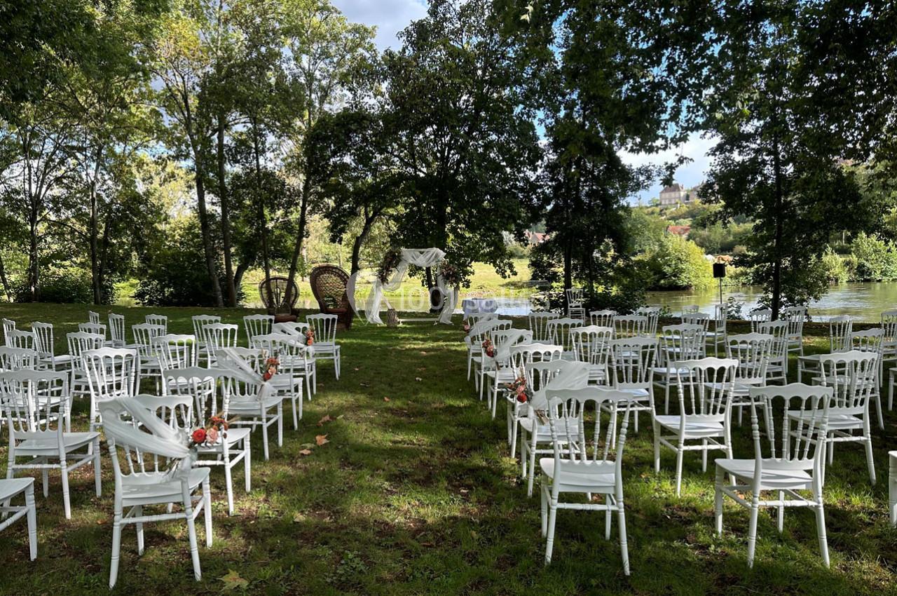 Chaises blanches disposées en rangées sur une pelouse, formant une allée centrale menant à une arche décorée pour une…