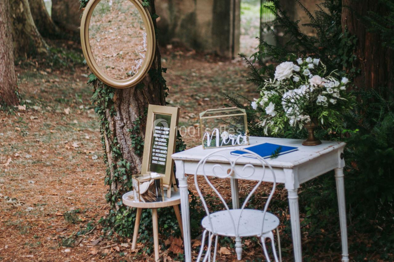 Table blanche décorée avec des fleurs, un miroir ovale fixé à un arbre et une chaise en métal dans un cadre boisé.