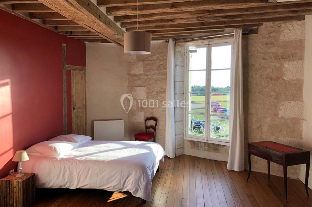 Chambre lumineuse avec lit double, mur rouge, poutres apparentes, parquet en bois et fenêtre donnant sur un paysage…