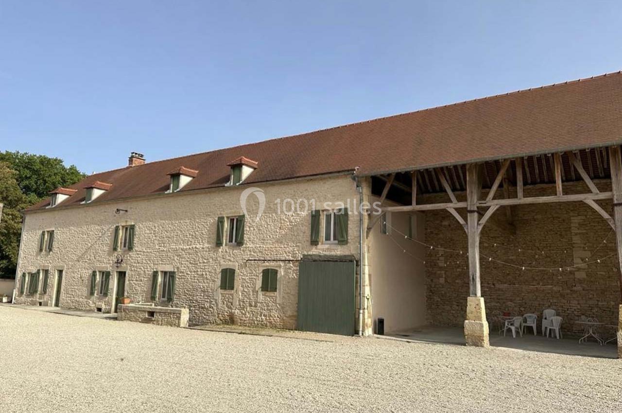 Bâtiment en pierre avec toit en tuiles, grandes fenêtres et une charpente ouverte, situé sur une cour gravillonnée.