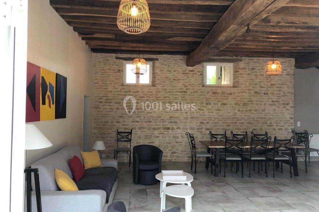 Salle de séjour avec murs en pierre, poutres apparentes, canapé gris, table à manger et chaises noires.