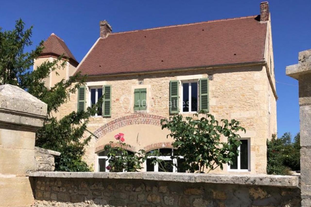 Façade d'une maison en pierre avec volets verts, toit en tuiles rouges et jardin fleuri, vue derrière un mur en pierre.