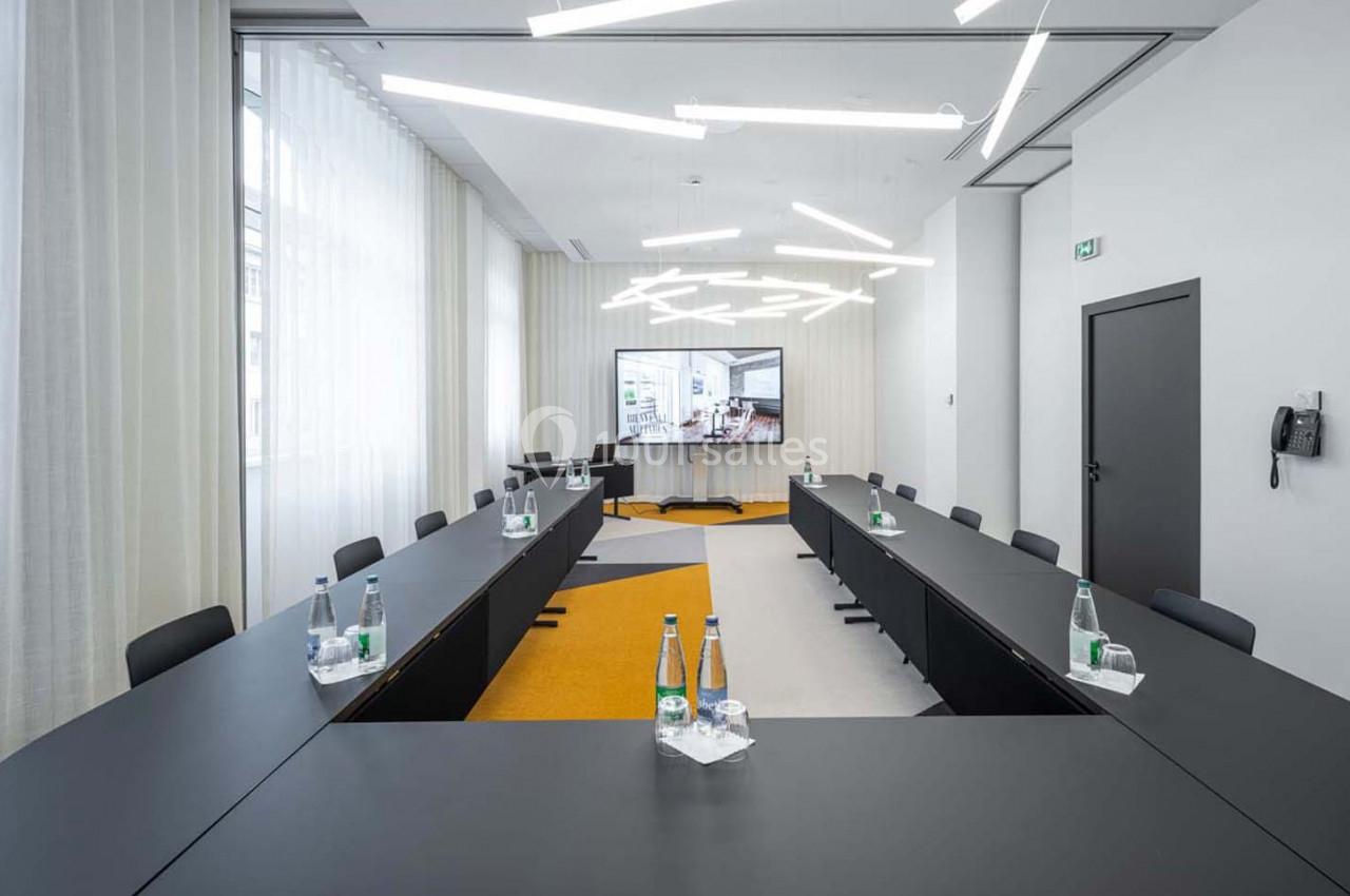 Salle de réunion moderne avec deux longues tables noires, bouteilles d'eau et écran affichant une présentation.