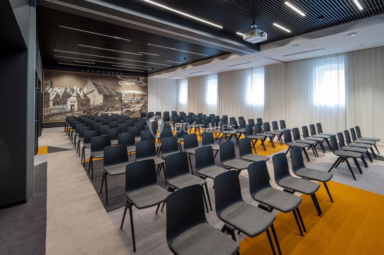 Salle de conférence moderne avec rangées de chaises noires, éclairage naturel et mural illustré en arrière-plan.