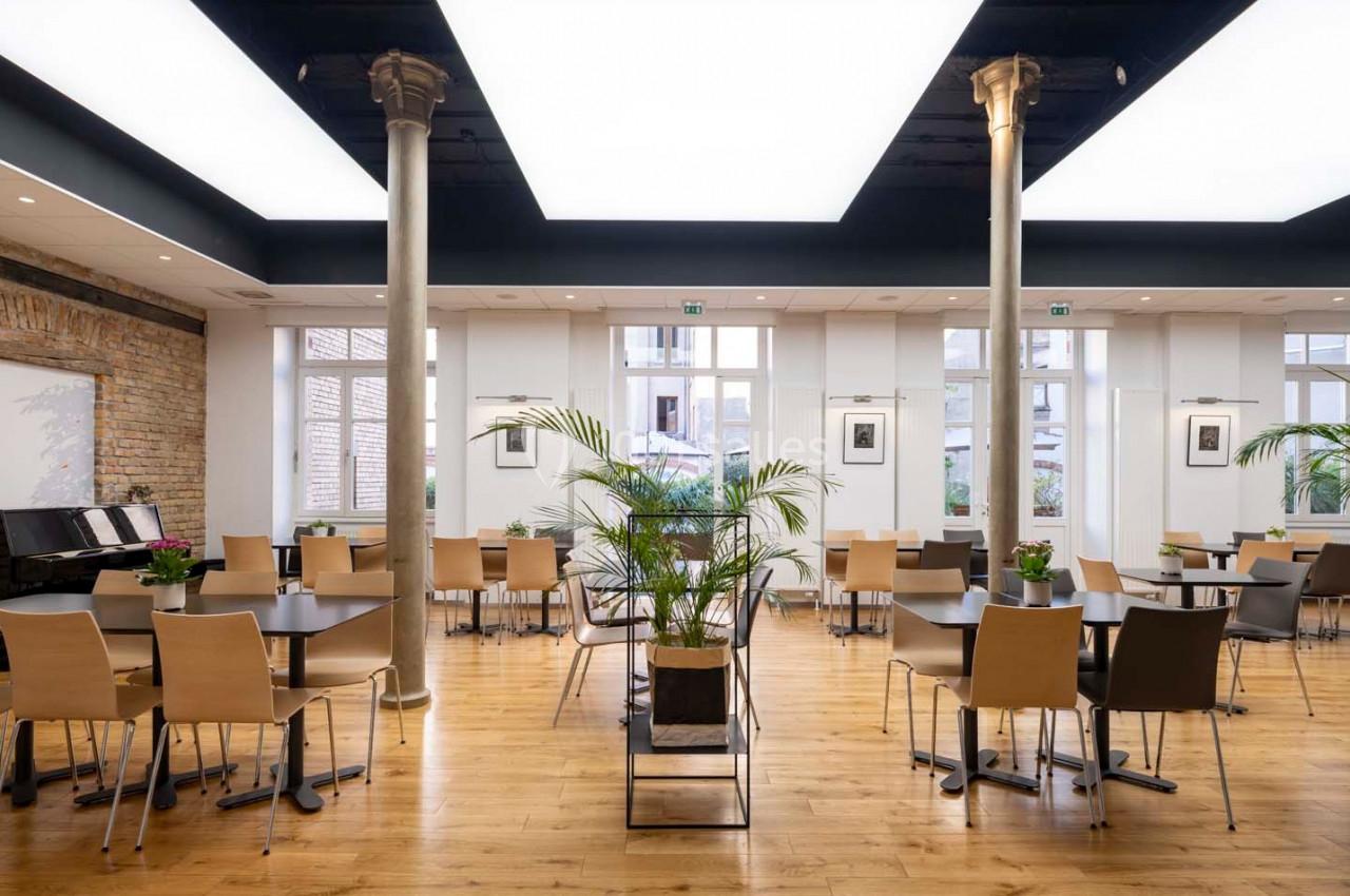 Salle lumineuse avec parquet, tables et chaises modernes, colonnes décoratives et plantes au centre.