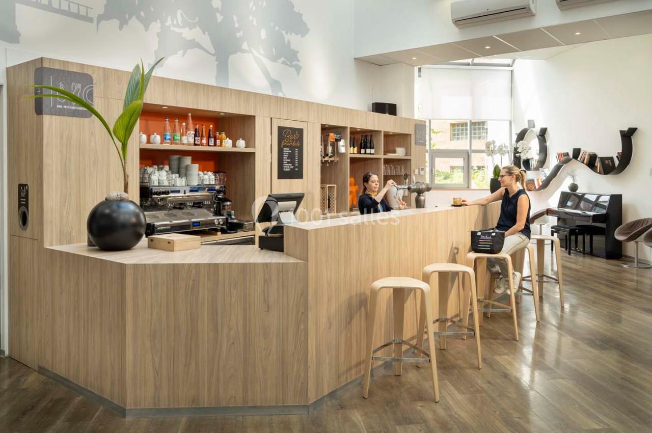 Un café moderne avec un comptoir en bois clair, des tabourets, une machine à café et des clients discutant.