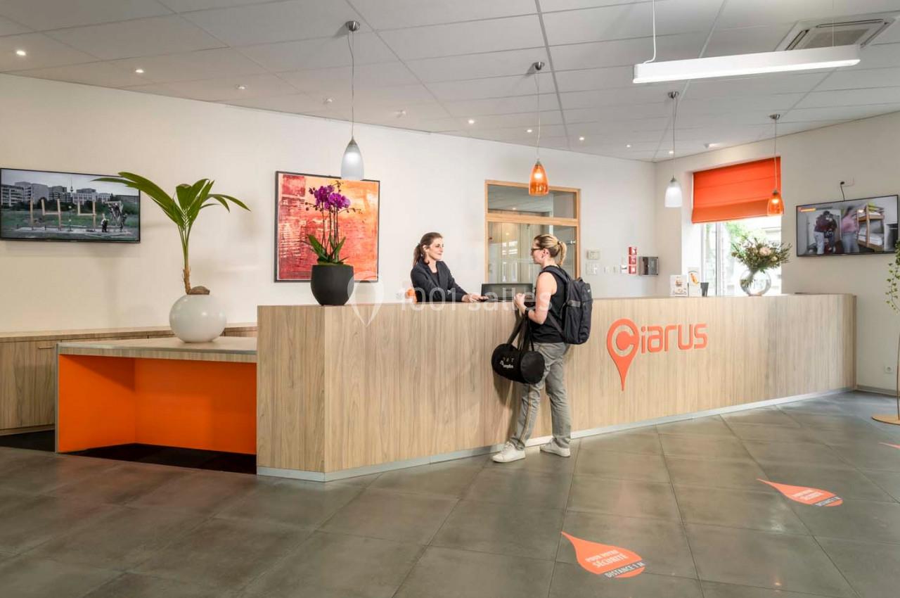 Accueil d'un établissement avec une réceptionniste échangeant avec un visiteur dans un espace lumineux et moderne.