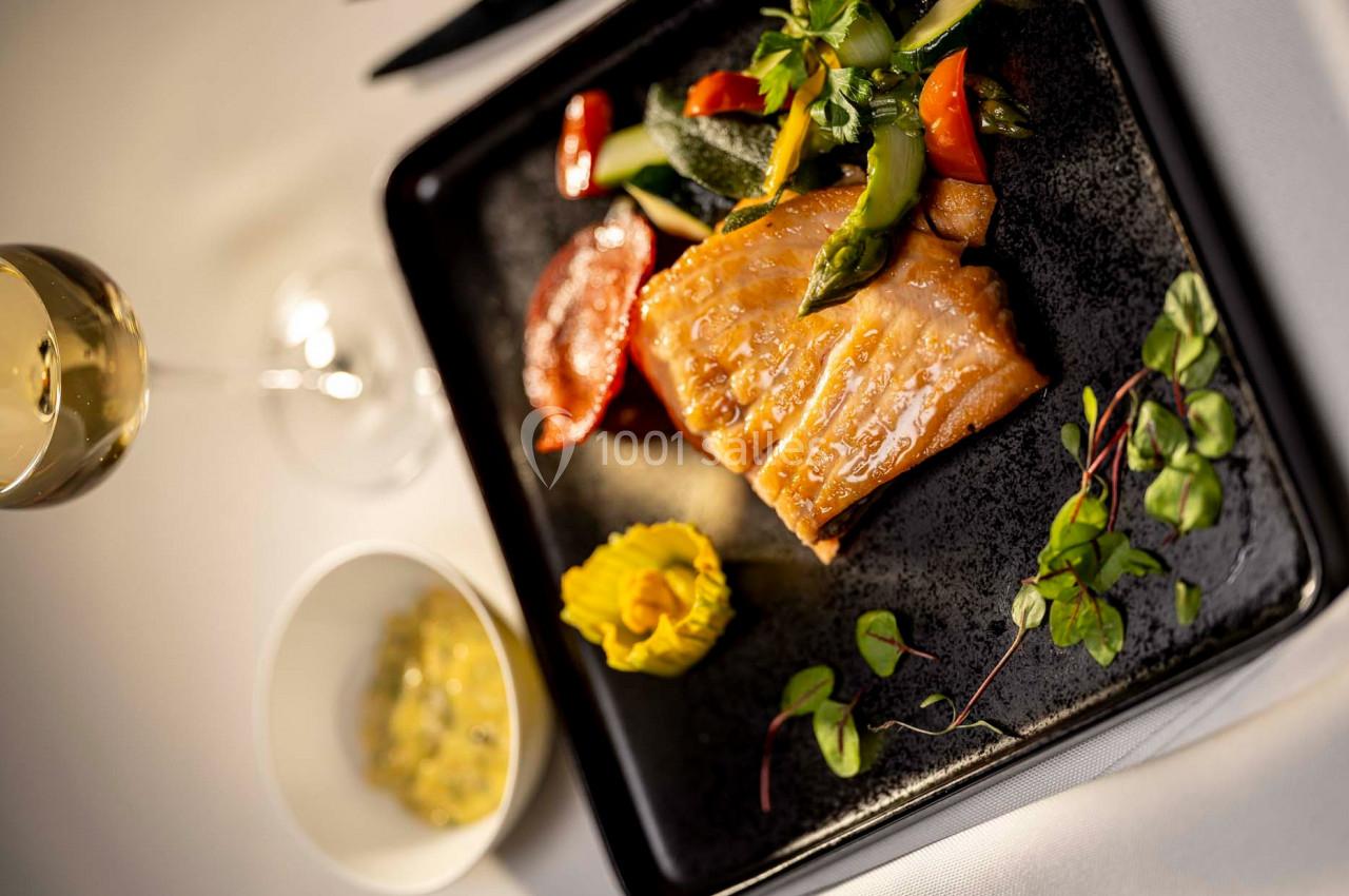 Assiette gastronomique avec filet de poisson grillé, légumes colorés, purée décorative et verre de vin en arrière-plan.