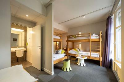 Chambre lumineuse avec un lit simple, des lits superposés en bois, un bureau et une fenêtre donnant sur des toits.