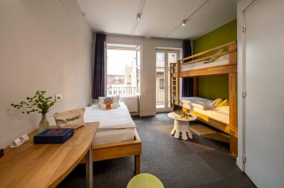 Chambre lumineuse avec un lit simple, des lits superposés en bois, un bureau et une fenêtre donnant sur des toits.