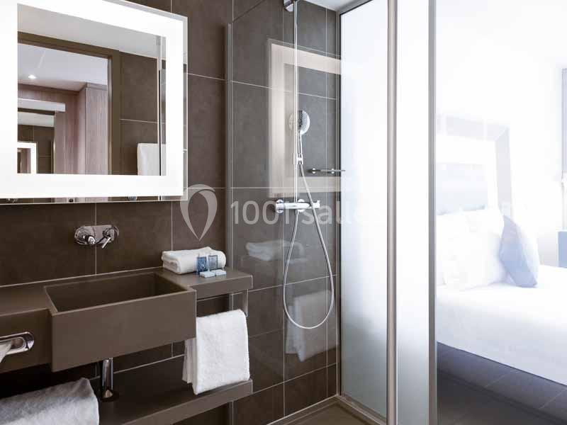 Salle de bain moderne avec douche vitrée, lavabo suspendu, miroir éclairé et serviettes pliées.