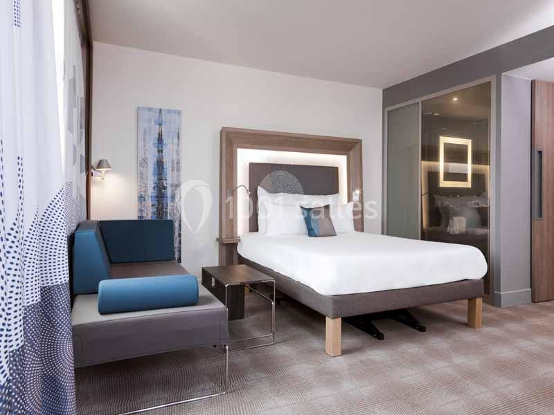 Chambre moderne avec lit double, canapé bleu, table basse et grande baie vitrée laissant entrer la lumière naturelle.